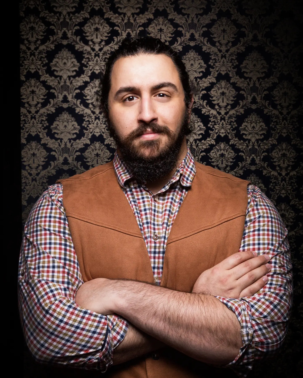 an actor with tied back long black hair  and a black beard wearing a tan vest and a blue, red, and white, checked shirt with sleeves rolled to the elbow stares  at the camera with one eyebrow raised and his arms crossed