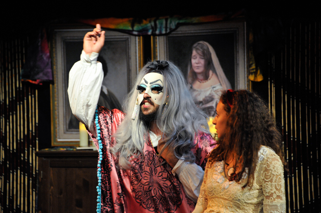 an actor with a blonde wig and a black beard wearing a pink and black floral dress and a white mask with makeup painted on and eyelashes attached stands with his arm up and his finger pointed to the ceiling dramatically facing out just behind another actor with long brown hair wearing a cream colored dress and a red headband who is turned to look at the first actor